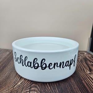 Op de afbeelding: Witte keramische voerbak met het woord "Schlabbernapf" in zwart schrift. De kom is gevuld met water en staat op een houten oppervlak. Een eenvoudig en functioneel ontwerp.