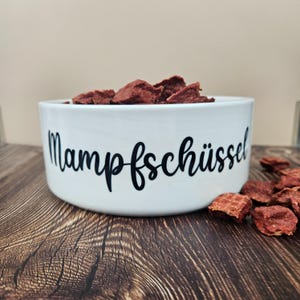Op de afbeelding: Een witte keramische voerbak voor huisdieren gevuld met roodbruine lekkernijen. De kom heeft het woord "Mampfsch&uuml;ssel" in zwart schrift. De kom staat op een houten oppervlak, met een paar lekkernijen verspreid. De achtergrond is een neutraal beige.