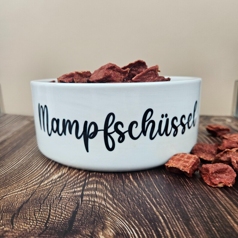 Op de afbeelding: Een witte keramische voerbak voor huisdieren gevuld met roodbruine lekkernijen. De kom heeft het woord "Mampfsch&uuml;ssel" in zwart schrift. De kom staat op een houten oppervlak, met een paar lekkernijen verspreid. De achtergrond is een neutraal beige.