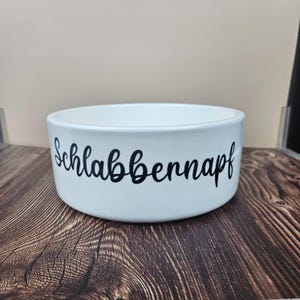 Op de afbeelding: Een witte keramische voerbak voor huisdieren met het woord "Schlabbernapf" in zwart schrift. De kom is rond met een brede opening en staat op een houten oppervlak. De kom is ontworpen voor dierenvoer of water.