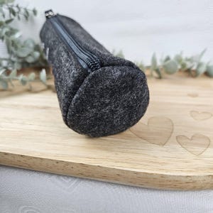 May include: A dark gray felt pencil case with a zipper closure. The cylindrical case is made of a textured fabric and is displayed on a wooden surface. The pencil case is designed to hold pens, pencils, and other stationery items.