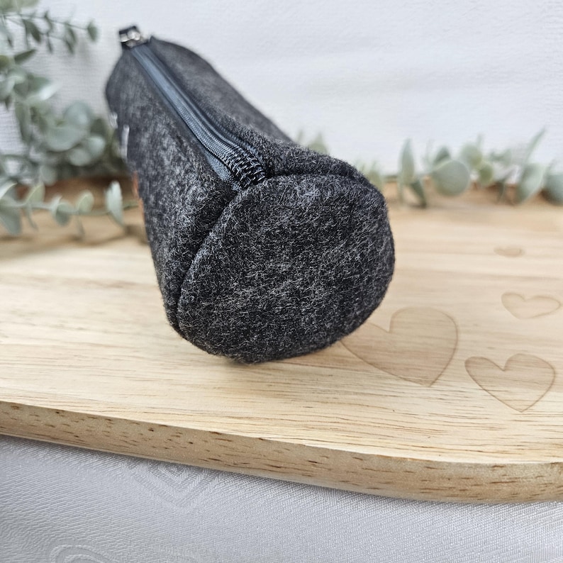 May include: A dark gray felt pencil case with a zipper closure. The cylindrical case is made of a textured fabric and is displayed on a wooden surface. The pencil case is designed to hold pens, pencils, and other stationery items.