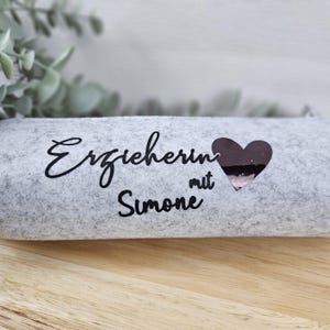 May include: A light gray felt pencil case with the words "Erzieherin mit Simone" in black script and a dark heart. The case is rectangular and rests on a wooden surface.