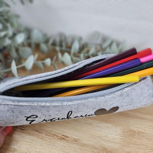 May include: A gray felt pencil case with a zipper, holding several colored pencils. The case has a heart and script text on the side. The pencils are yellow, red, purple, and other colors.