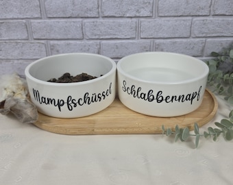 May include: Two white ceramic pet bowls placed on an oval wooden tray. The left bowl contains dark food and is labeled with the black text Mampfschüssel. The right bowl is filled with water and is labeled with the black text Schlabbernapf.