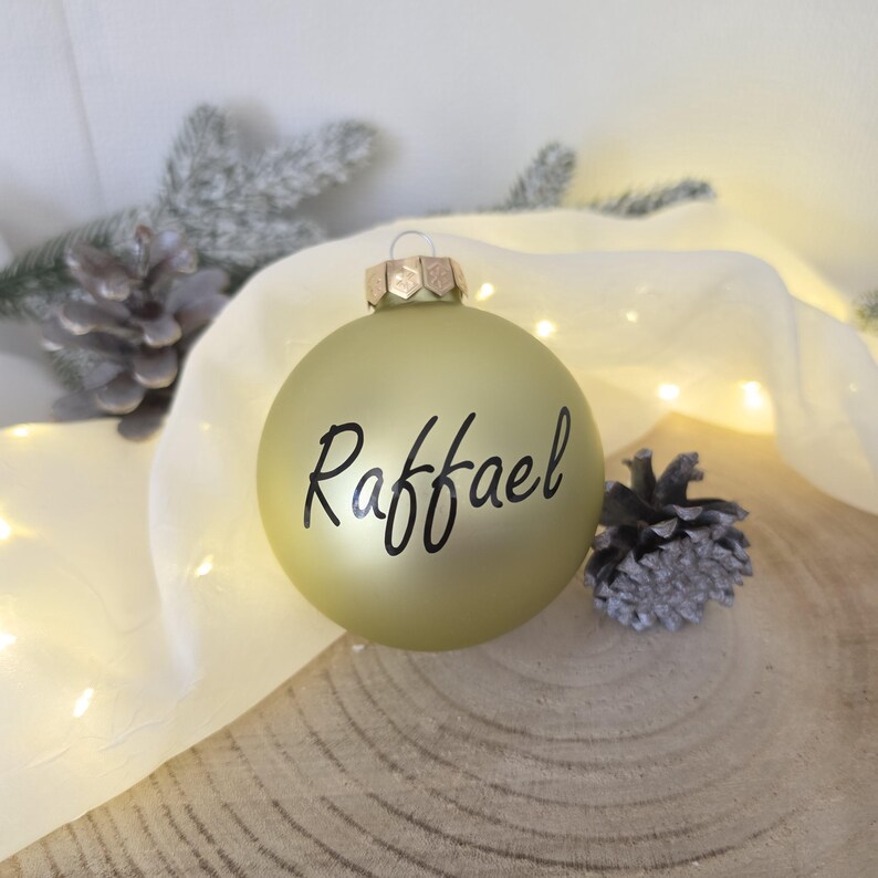 May include: A matte gold spherical Christmas ornament featuring the name Raffael written in black cursive script. The ornament has a metallic silver cap at the top and is designed for hanging on a tree or as a decorative holiday display piece.