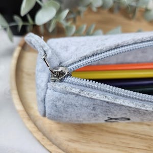 May include: A light gray felt pencil case with a silver zipper, partially open to reveal colorful pencils inside. The pencil case has a small fish design on the side. The case is on a wooden tray.