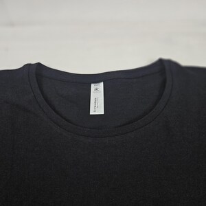 K&ouml;nnte beinhalten: Ein einfarbiges schwarzes T-Shirt mit Rundhalsausschnitt. Ein wei&szlig;es, rechteckiges Stoffetikett ist an der Innenseite des Kragens befestigt und zeigt den Text B&C Collection sowie die Worte for women in schwarzer Tinte auf wei&szlig;em Grund.