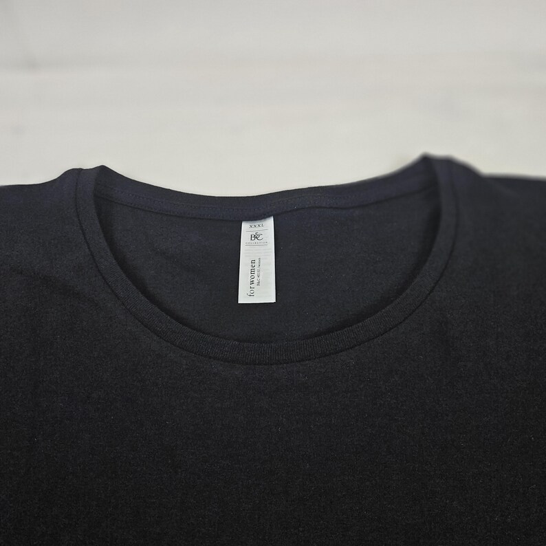 K&ouml;nnte beinhalten: Ein einfarbiges schwarzes T-Shirt mit Rundhalsausschnitt. Ein wei&szlig;es, rechteckiges Stoffetikett ist an der Innenseite des Kragens befestigt und zeigt den Text B&C Collection sowie die Worte for women in schwarzer Tinte auf wei&szlig;em Grund.