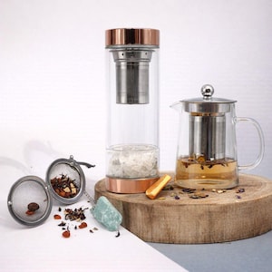May include: A tall glass tea infuser bottle with a copper-toned lid and base, a glass teapot with a stainless steel infuser containing amber liquid, and a round metal mesh tea ball infuser with loose leaf tea and a light green crystal charm.