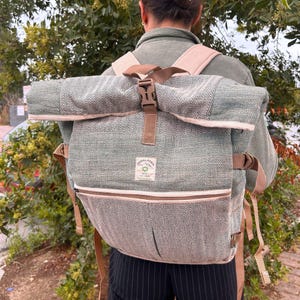 May include: A roll-top backpack featuring a sage green and beige herringbone pattern. The bag includes a brown plastic buckle closure, adjustable shoulder straps, and a front zippered pocket. A rectangular label on the front reads Himalayan.