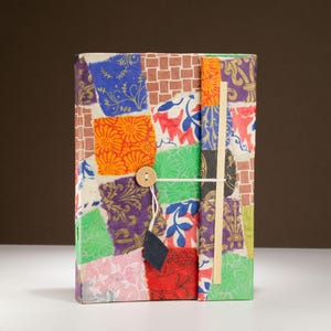 Peut inclure: Un journal rectangulaire recouvert d'un patchwork de papier coloré présentant divers motifs floraux, géométriques et de vignes dans des tons orange, violet, vert, rouge et bleu. Une fermeture à bouton en bois avec un cordon blanc sécurise la couverture.