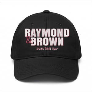 May include: Black baseball cap with the text "RAYMOND & BROWN" in white and pink lettering. Below the text is "2026 R&B Tour" in a smaller pink font. The cap has a curved brim and a button on top.