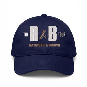 May include: A navy blue baseball cap with the text "THE R&B TOUR" in white and brown. The words "RAYMOND & BROWN" are printed below the main text. The cap has a curved brim and a button on top.