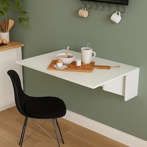 May include: A white, wall-mounted folding table with a black chair. A wooden cutting board holds a bowl of cereal, an egg, and a mug. A black shelf above holds spices, plants, and mugs. The table is approximately 40 inches long.