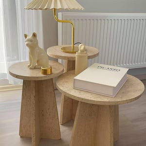 Puede incluir: Un conjunto de tres mesas auxiliares beige con superficie texturizada. Una lámpara dorada con pantalla plisada se encuentra sobre una mesa. También se exhiben una figura de gato de cerámica, un pequeño recipiente dorado, una botella y un libro titulado "PICASSO".