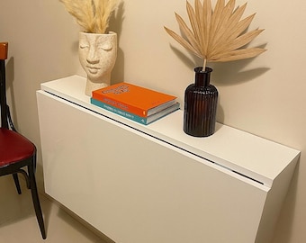 Könnte beinhalten: Ein weißer, an der Wand befestigter Klapptisch mit minimalistischem Design. Er hat eine schwarze Zierleiste und ist mit dekorativen Gegenständen geschmückt: einer cremefarbenen Gesichtsvase mit Pampasgras, zwei Büchern und einer dunkelbraunen Vase mit einem getrockneten Palmblatt.