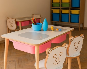 May include: A children's playroom with a white table and two chairs. The table has a pink storage bin and a gray inset. The chairs have bear-shaped backs. A blue vase and bowl sit on the table. A wall decoration and storage shelves are in the background.