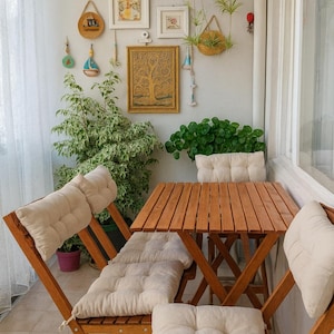 May include: A cozy balcony scene featuring a wooden folding table and chairs with cream-colored cushions. The walls are adorned with framed artwork and decorative hanging items. Potted plants and hanging baskets add a touch of greenery to the space.