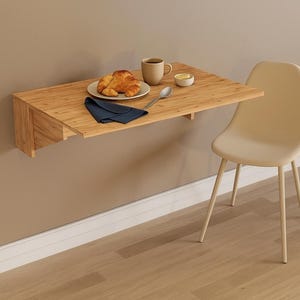 Peut inclure: Table pliante en bois clair fixée au mur avec croissant, café et beurre. Une chaise beige est à côté de la table. Un support en métal noir avec des tasses beiges est au-dessus. La table mesure environ 76 cm de long.