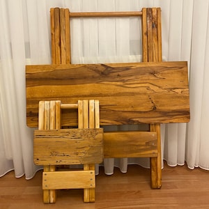 Puede incluir: Un juego de dos mesas plegables de madera. La mesa más grande tiene una tapa rectangular y un asa en la parte superior. La mesa más pequeña es una versión en miniatura de la más grande. Ambas mesas son de madera marrón claro con vetas visibles.
