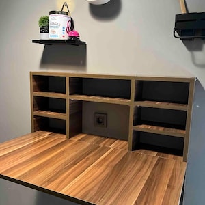 Könnte beinhalten: Ein wandmontierter Schreibtisch aus Holz mit einer herunterklappbaren Arbeitsfläche. Der Schreibtisch hat eingebaute Regale und ein zentrales Fach. Das Holz hat eine warme, natürliche Maserung. Eine weiße Ukulele hängt an der Wand über dem Schreibtisch.