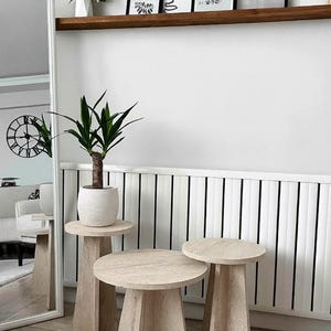 Puede incluir: Un conjunto de tres mesas auxiliares de piedra beige con tapa redonda y base geométrica. Una planta en maceta se encuentra sobre la mesa más alta. Las mesas están dispuestas frente a una pared blanca con rayas verticales negras.