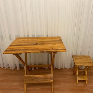 Puede incluir: Una mesa plegable de madera y un taburete a juego. La mesa tiene una tapa rectangular y patas en ángulo. El taburete es una versión más pequeña de la mesa. Ambos son de madera marrón claro con vetas visibles.