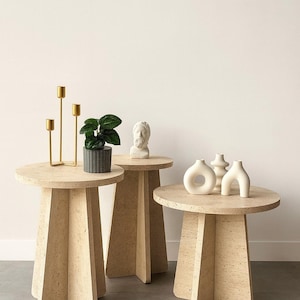 Puede incluir: Tres mesas auxiliares beige con tapas redondas y bases geométricas. Las mesas están decoradas con un candelabro dorado, una pequeña planta en maceta, un busto y jarrones de cerámica blanca. Las mesas están hechas de un material claro y texturizado.