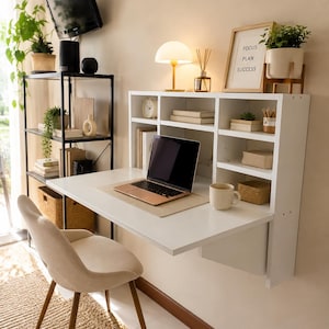 May include: A white wall-mounted desk with integrated shelving units, featuring a laptop, a beige mug, and a framed sign that reads FOCUS PLAN SUCCESS. A beige upholstered chair with wooden legs is positioned in front of the desk.