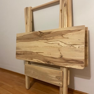 May include: A light-coloured, wooden folding table with a natural wood grain pattern. The table has a rectangular top and a handle at the top for easy carrying. The legs are folded for storage.