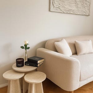 May include: A light-toned living room with a beige sofa and cushions, three small round side tables, and textured wall art. A candle, a rose in a vase, and books are on the tables. The tables are made of a light-colored material.