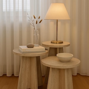 Puede incluir: Conjunto de tres mesas auxiliares beige. Una tiene una lámpara, un libro con la palabra «Interiors» y una piedra decorativa. Otra mesa tiene un cuenco blanco. La lámpara tiene una pantalla beige y una base dorada.