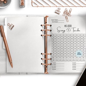 May include: A white planner with a gold binder ring and a savings tracker page. The page has a grid for tracking $5,000 in savings with a start date and end date section. The text "$5,000 Savings Tracker" is at the top of the page.