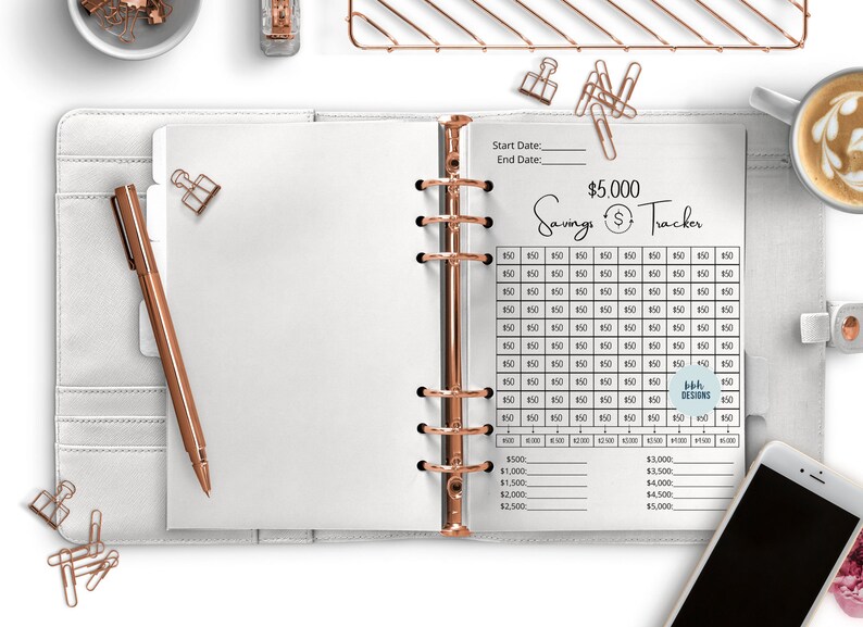 May include: A white planner with a gold binder ring and a savings tracker page. The page has a grid for tracking $5,000 in savings with a start date and end date section. The text "$5,000 Savings Tracker" is at the top of the page.