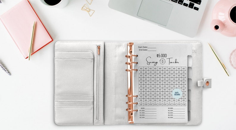 May include: A white binder with a clear plastic sheet inside. The sheet has a savings tracker with a goal of $5,000. The tracker is divided into 100 squares, each representing $50. The sheet also has a section for recording start and end dates.