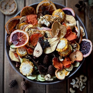 May include: A bowl filled with an assortment of dried fruits and vegetables, including orange slices, apple slices, and corn. The bowl also contains cashews, pine cones, and other decorative elements. The arrangement is set on a wooden surface.