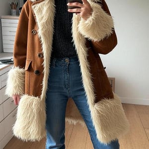 May include: A brown suede coat with cream faux fur trim on the collar, cuffs, and front. The coat features a double-breasted design with large buttons. It is worn with blue denim jeans.
