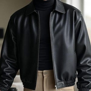 Könnte beinhalten: Schwarze Lederjacke mit Kragen und silbernem Reißverschluss. Die Jacke hat gerippte Bündchen und einen gerippten Saum. Die Person trägt einen schwarzen Rollkragenpullover und eine beige Hose. Eine Kaffeetasse ist sichtbar.