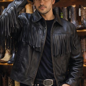 Puede incluir: Chaqueta de cuero negro con flecos en las mangas y el pecho. La chaqueta se lleva con un sombrero de vaquero negro, vaqueros negros y un cinturón con hebilla decorativa. Botas de vaquero visibles en el fondo.