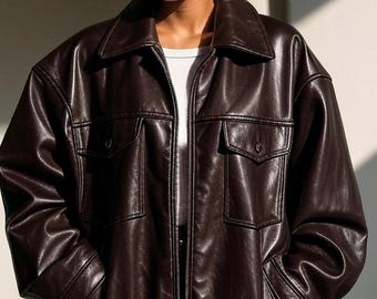 May include: A dark brown leather jacket with a classic collar, two chest pockets, and a front zipper closure. The jacket has a slightly oversized fit and buttoned cuffs. The model is wearing a white undershirt.