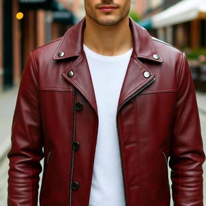 Op de afbeelding: Bordeaux leren jas met een klassiek design. De jas heeft een reverskraag, een ritssluiting en knopen. Het model draagt er een wit t-shirt onder. De achtergrond is een vage straatscène.