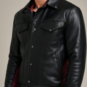 Könnte beinhalten: Schwarze Lederjacke mit klassischem Kragen, zwei Brusttaschen und Knopfverschluss. Die Jacke hat eine taillierte Passform und ein rotes Steppfutter, das an den Manschetten und im Inneren sichtbar ist. Ein stilvolles Oberbekleidungsstück.