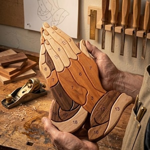 May include: A wooden puzzle in the shape of praying hands, featuring various shades of brown and tan wood. The puzzle is held by a person. Numbers are visible on the puzzle pieces. A small hand plane and woodworking tools are in the background.