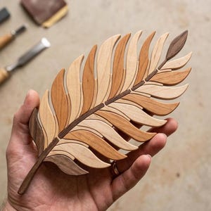 May include: A handcrafted wooden leaf sculpture with a detailed design. The leaf is composed of various shades of wood, including light and dark browns, and is held in a hand. The sculpture showcases a natural, organic theme.