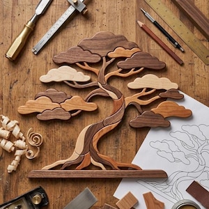 May include: A wooden bonsai tree sculpture crafted from various shades of light and dark wood, featuring layered cloud-like foliage and a curved trunk, mounted on a rectangular base, surrounded by woodworking tools and a paper sketch.