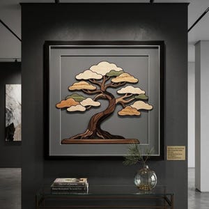 May include: A square wall art piece featuring a layered wooden bonsai tree with a dark brown trunk and light wood foliage in a black frame. The tree is set against a gray background. A small gold plaque with text is mounted on the wall nearby.