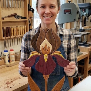 May include: A handcrafted wooden wall art piece shaped like an iris flower, featuring segmented petals in various wood tones including dark brown, light tan, purple, and yellow. Each wooden segment is numbered with black digits from 4 to 17.