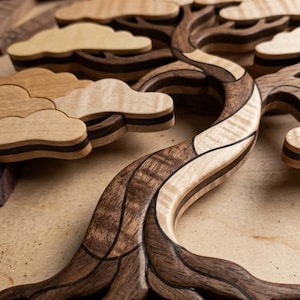 May include: A wooden wall art piece shaped like a bonsai tree, constructed from contrasting light and dark wood segments. The design features layered, cloud-like foliage sections and a curved, textured trunk with visible wood grain patterns.