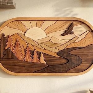 May include: A wooden wall art piece depicting a mountain landscape with a rising sun and a flying bird. The artwork features a river, trees, and mountains, all crafted from various shades of wood. The piece is framed in a light brown, oval-shaped border.
